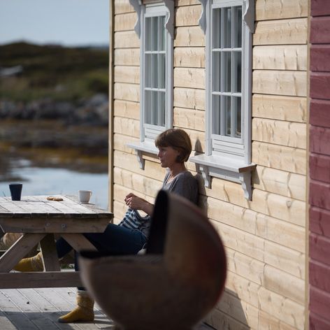 Kvinne sitter på benk utenfor rorbu og strkker og koser seg med en kopp kaffe i solsteiken
